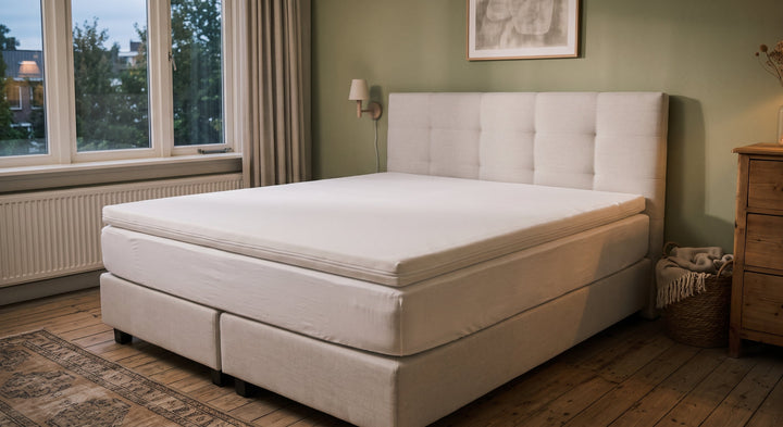 Een beige tweepersoons boxspring met een gecapitonneerd hoofdbord en een topper in een rustige slaapkamer.