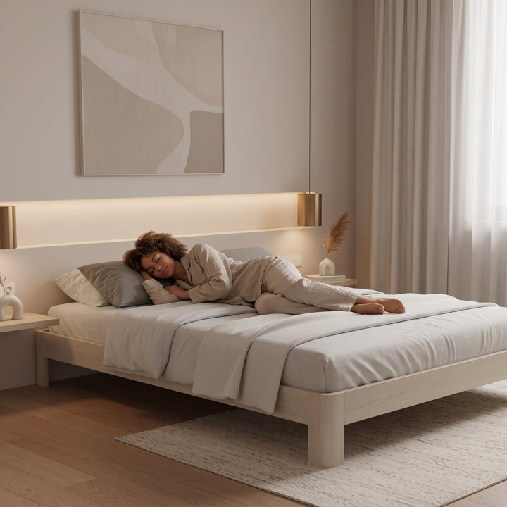 Vrouw slaapt op zij in serene, minimalistische slaapkamer. Neutrale tinten, licht hout, abstracte kunst. Daglicht door groot raam.